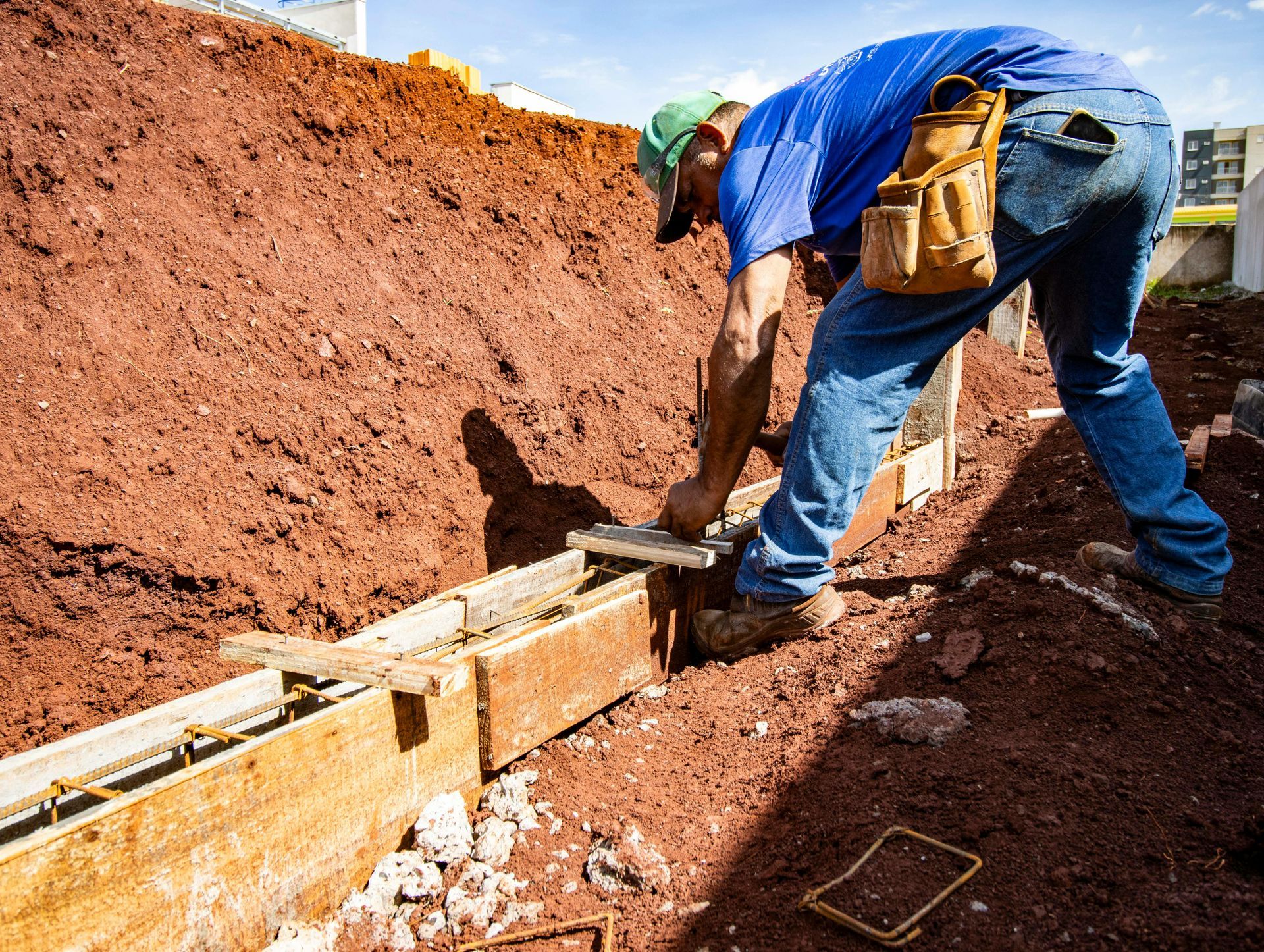 A construction worker wearing a blue shirt and tool belt works on a wooden foundation form in a trench of red soil.