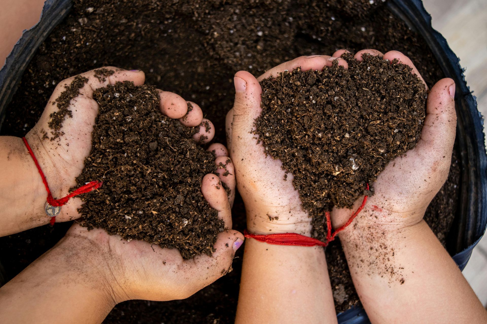 Two pairs of cupped hands, each wearing a red string bracelet, hold a handful of rich, dark potting soil over a container.