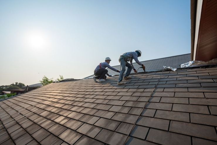 Two roofers installing shingles on a house roof under a bright sky.