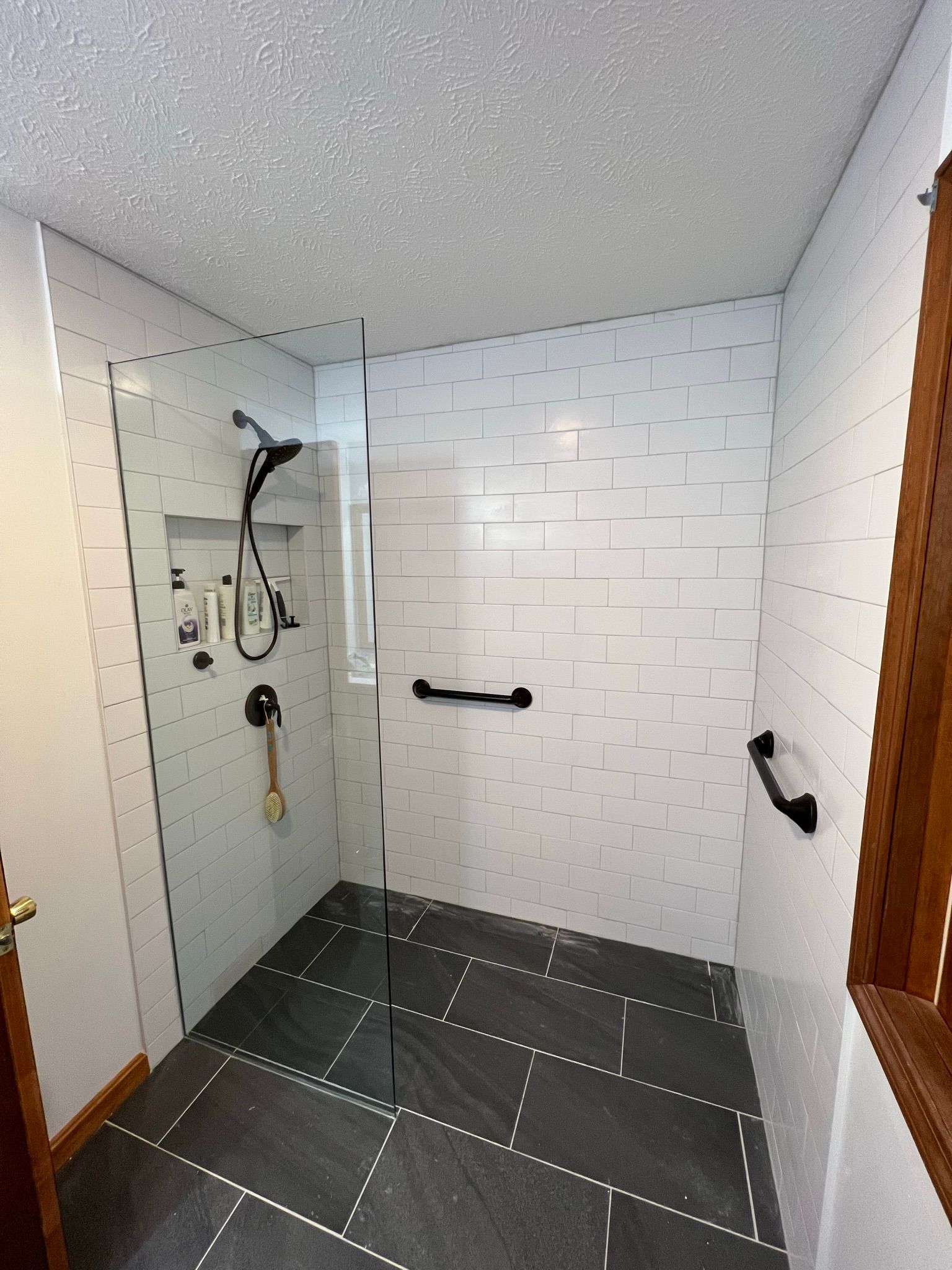 A modern, accessible shower with white tile walls, a black tiled floor, and black grab bars.