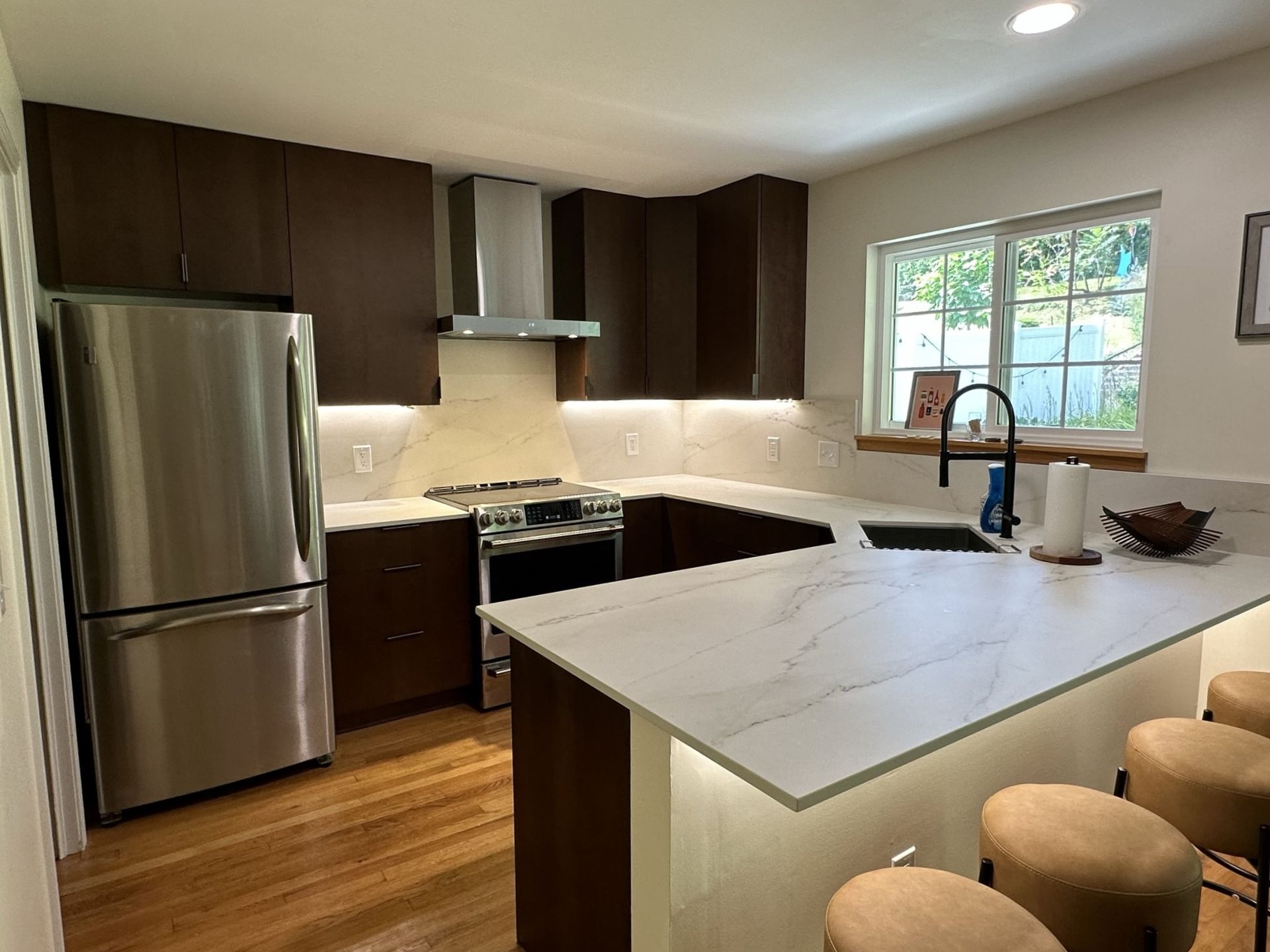Modern kitchen with dark brown cabinets, stainless steel appliances, and a white countertop. A window provides natural light.