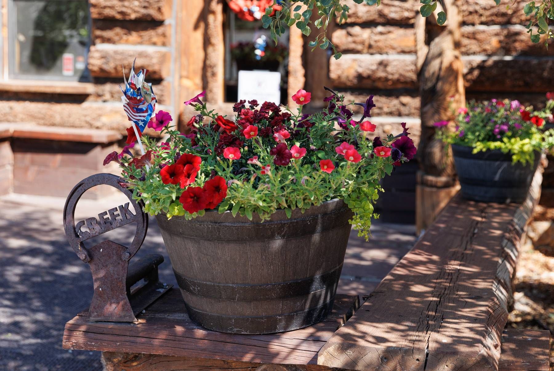 A barrel filled with flowers sits on a wooden table in front of a log cabin.