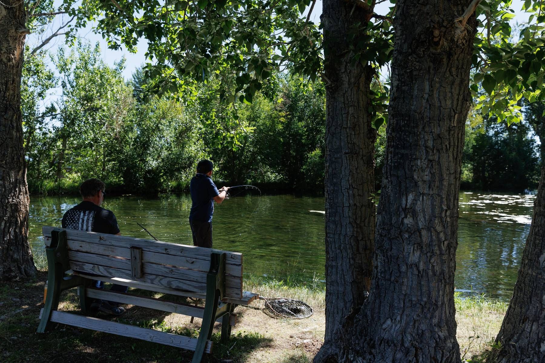 A man is fishing in a pond while another man sits on a bench.