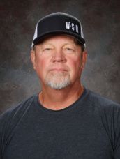 A man wearing a hat and a green shirt is standing in front of a green background.