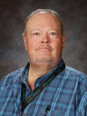 A man in a blue plaid shirt is standing in front of a green background.