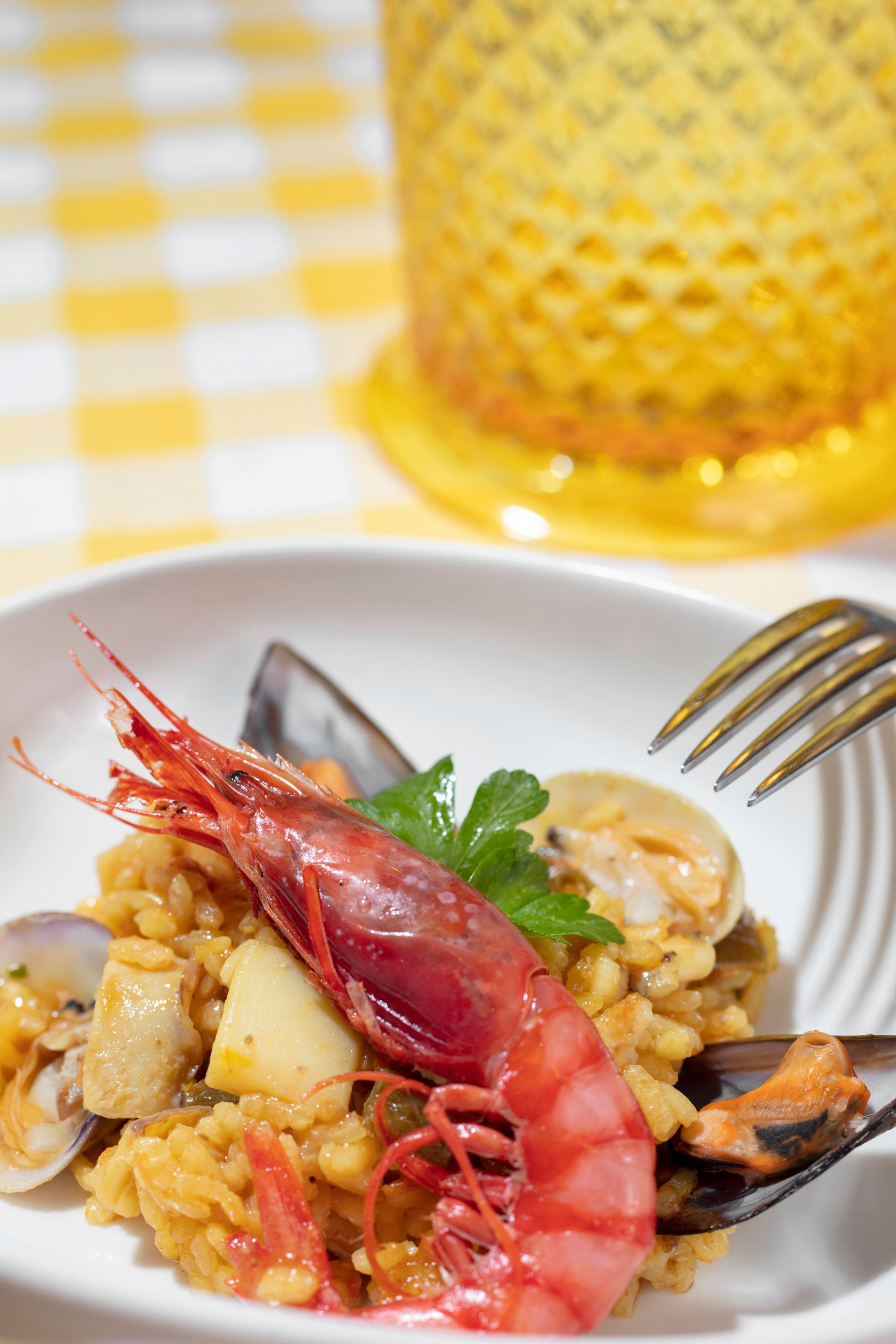 Paella con gamberi e frutti di mare, servita su un piatto bianco con una forchetta, tovaglia a quadri gialli.