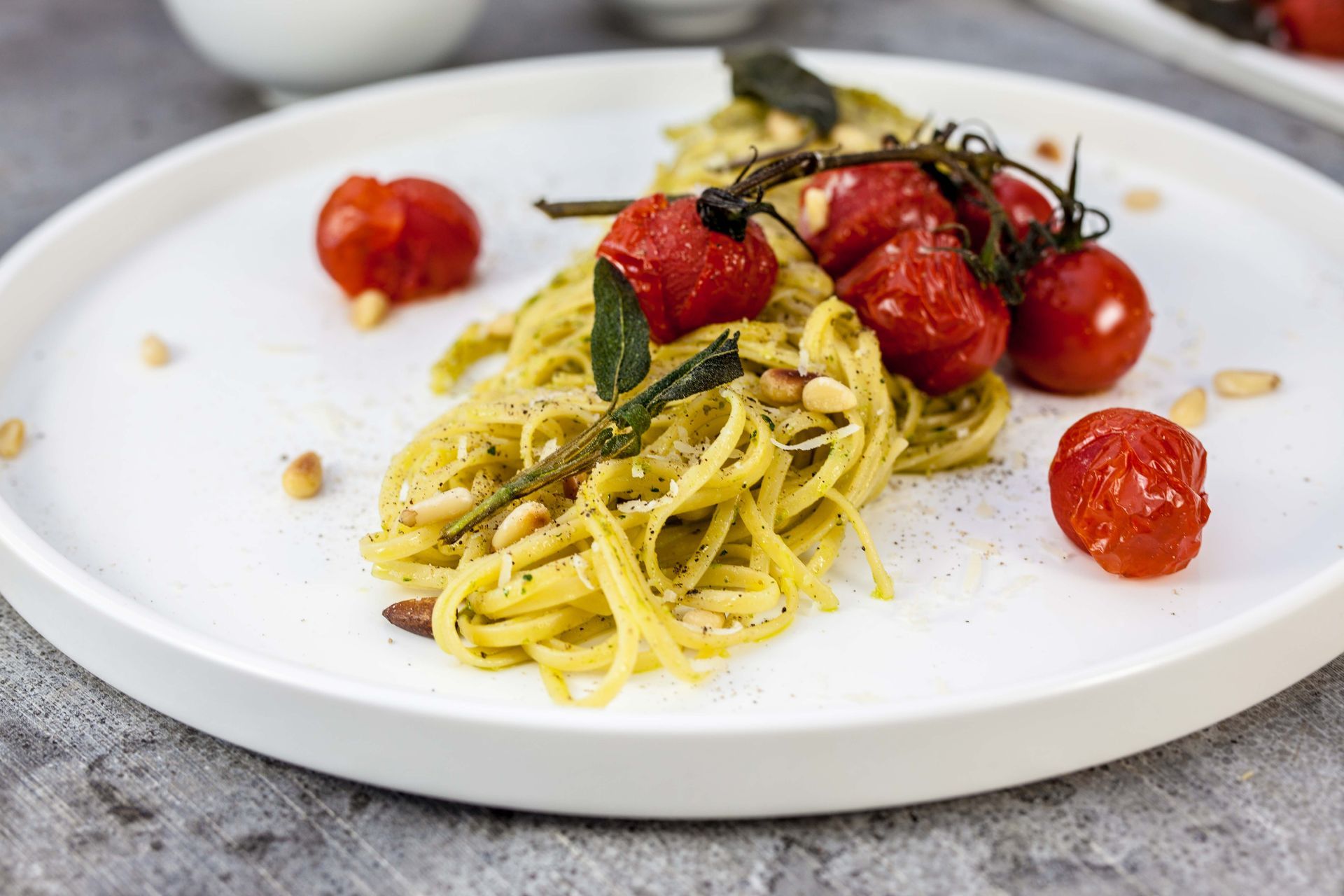 Linguine con pesto, pomodori arrostiti, pinoli e salvia fritta su un piatto bianco.