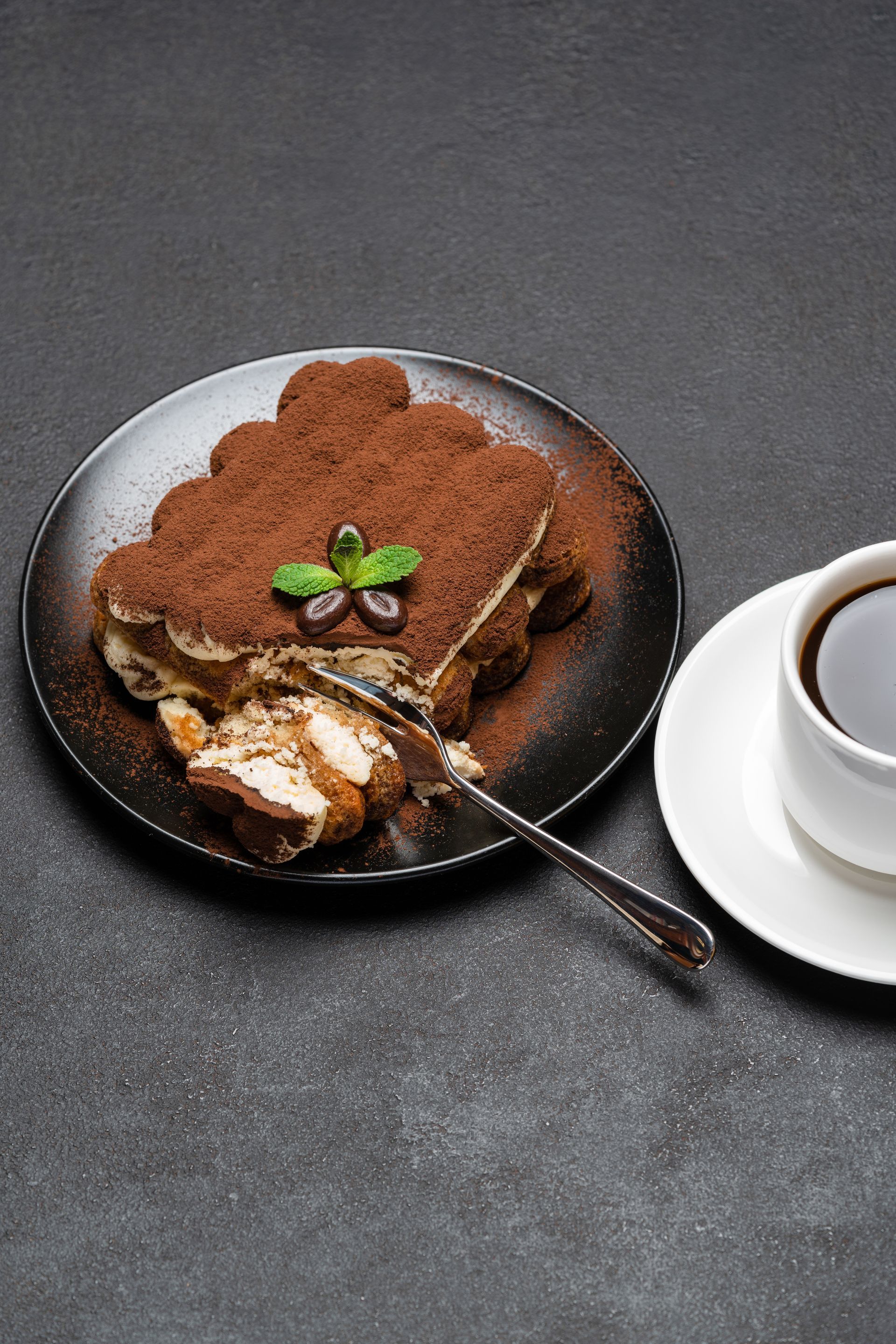 Tiramisù servito su un piatto scuro, spolverato con cacao in polvere e con una tazza di caffè a lato.