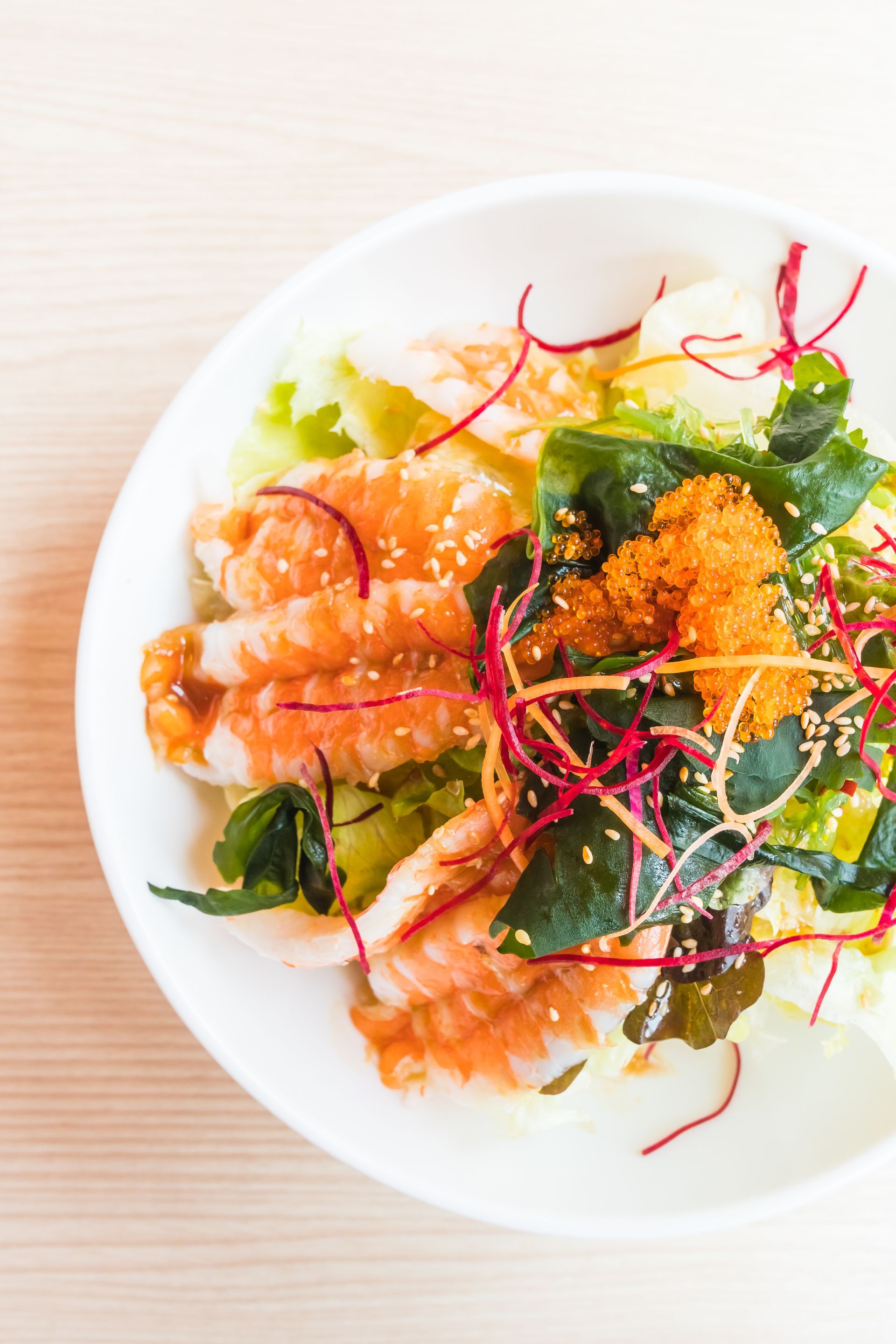 Ciotola bianca di insalata di gamberi con alghe, fili rossi e uova d'arancia su una superficie di legno chiaro.