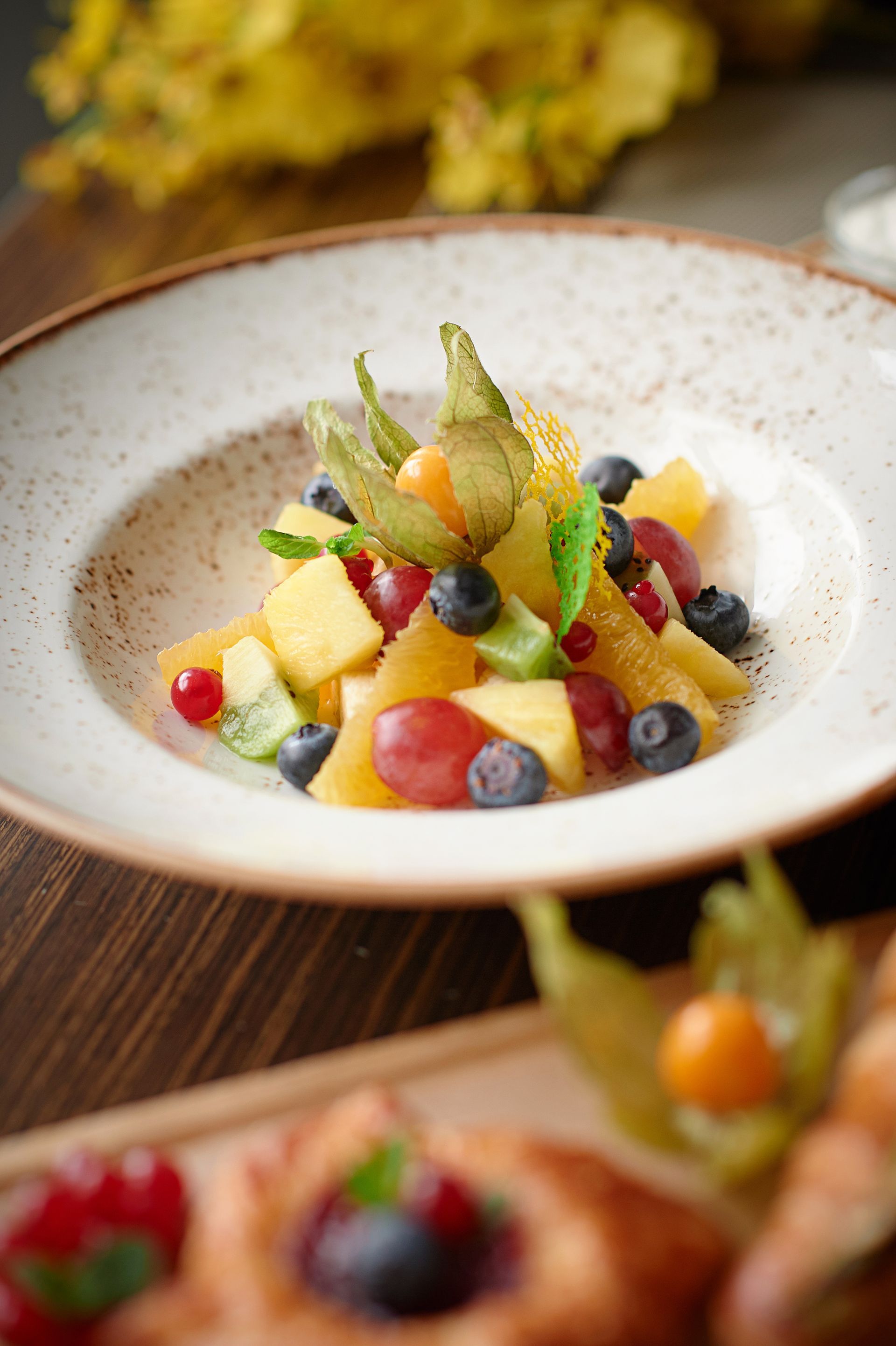 Macedonia di frutta con ananas, frutti di bosco e alchechengi in una ciotola bianca, primo piano.