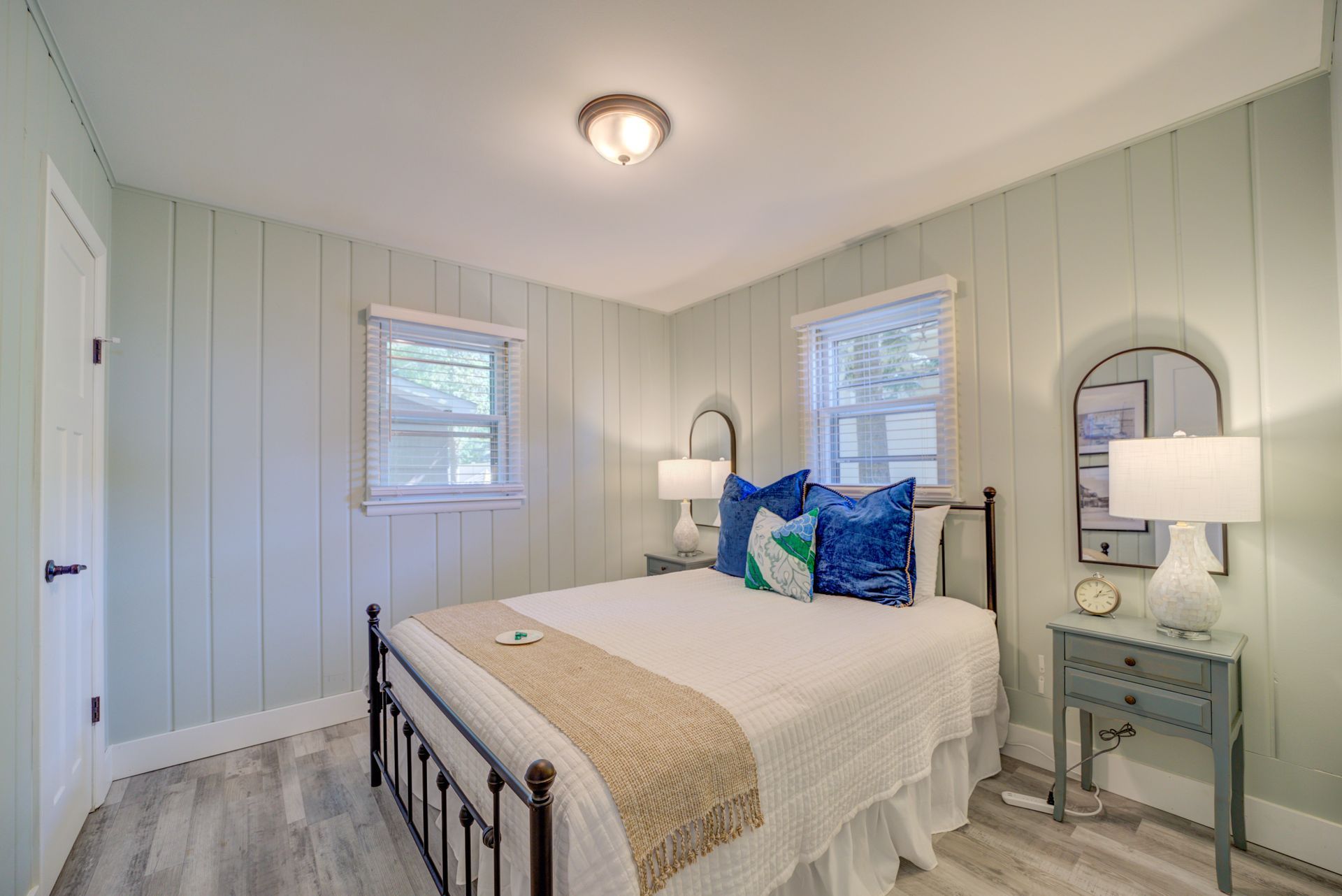 A bedroom with a bed , nightstand , lamps and two windows.
