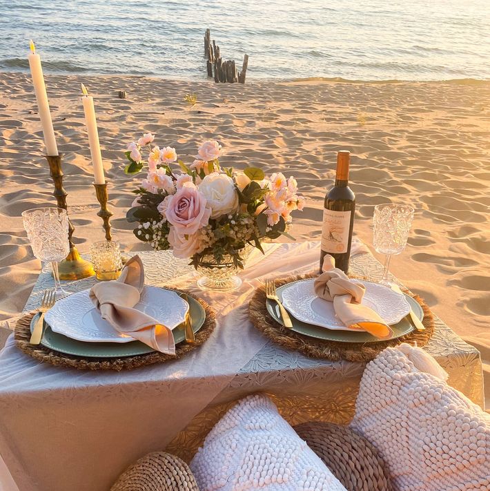 A table with plates , candles , and a bottle of wine on the beach.