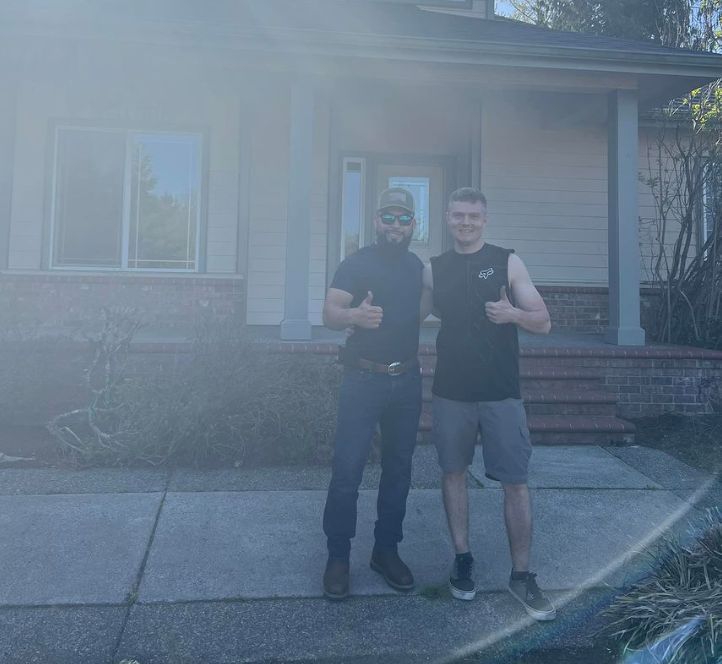 Two men are standing in front of a house giving thumbs up.