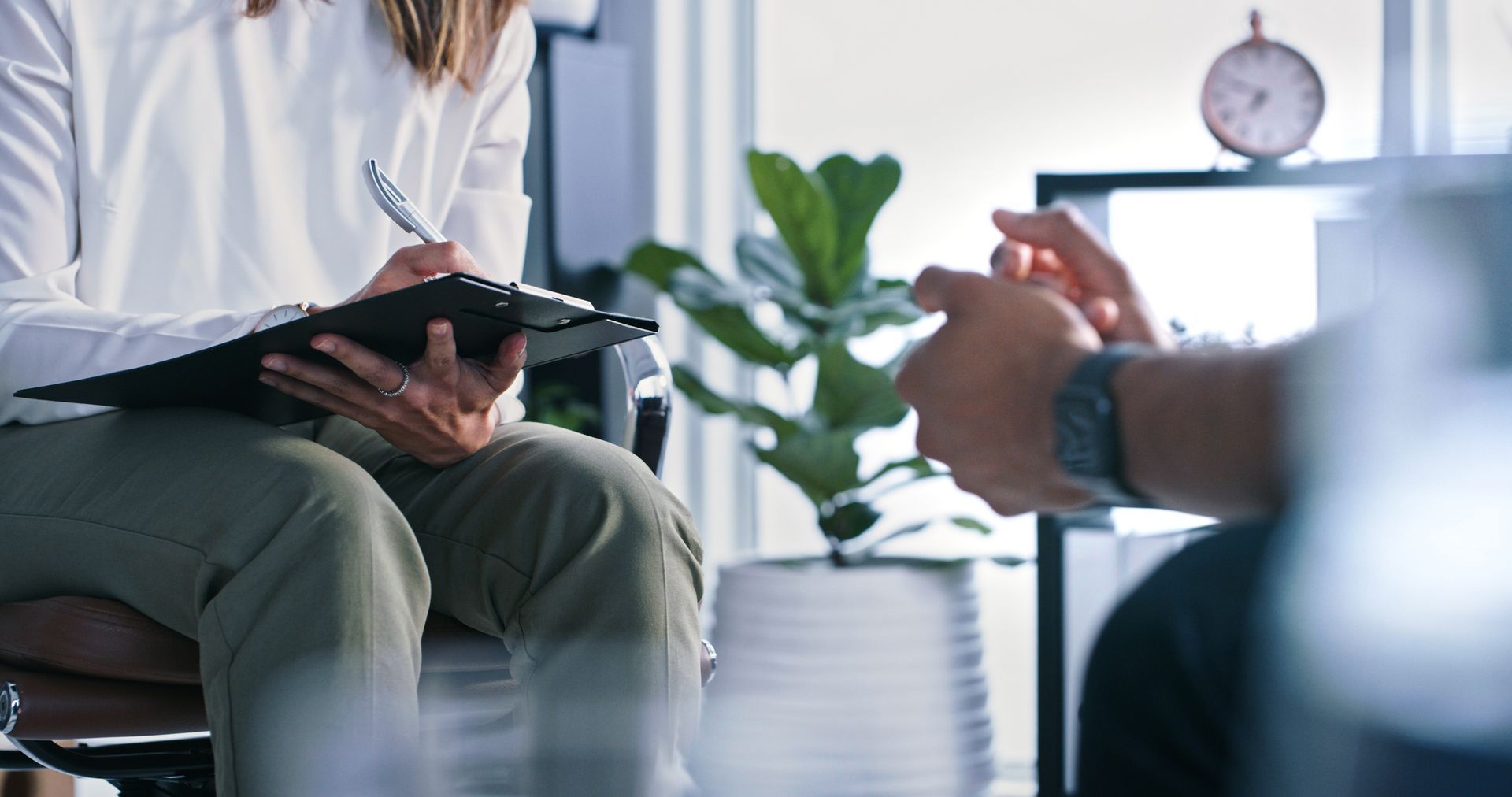 A woman is sitting in a chair talking to a man while holding a clipboard.