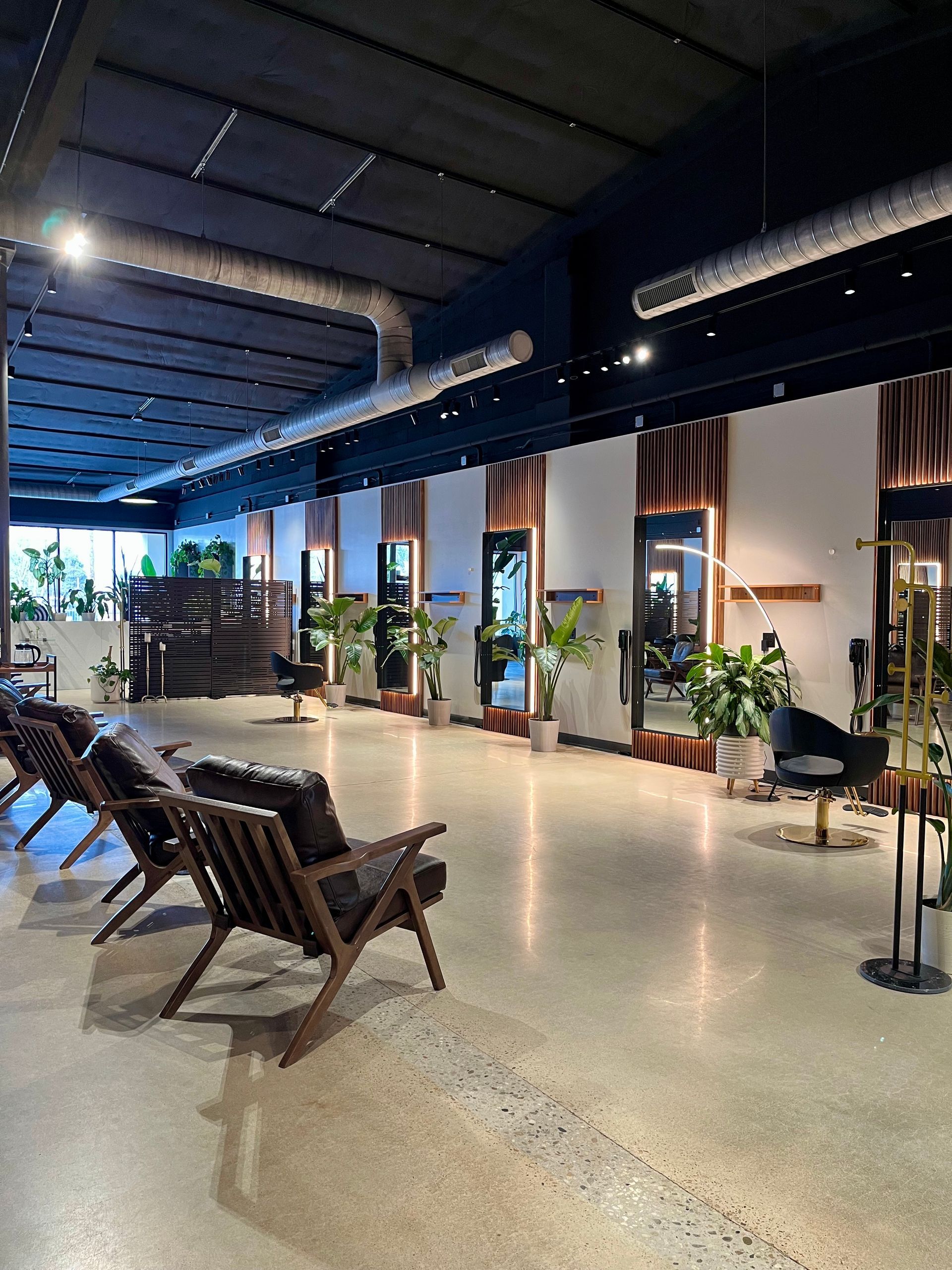 Salon interior with large mirrors, plants, seating, and exposed ducts.  Modern design with wood and black accents.