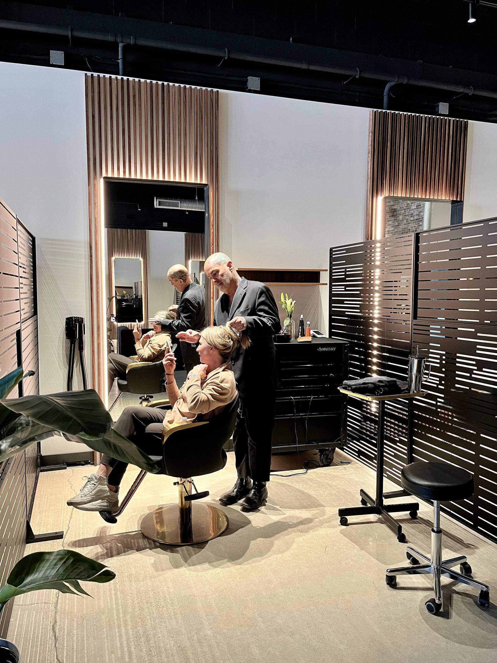 Hair salon interior with stylists and clients. Features include mirrors, plants, and dividers; a phone number and Instagram handle are displayed.