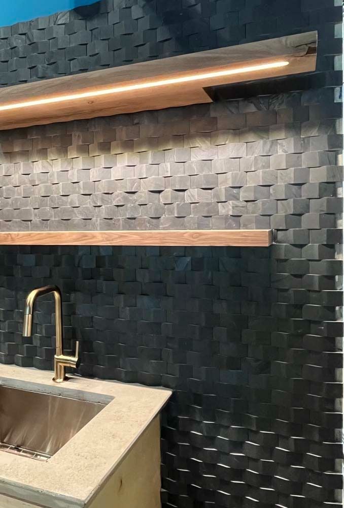 Kitchen sink with gold faucet, set against textured dark tile wall, wooden shelves, and strip lighting.