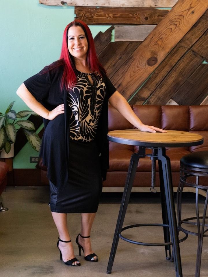 Woman with red hair, in a black dress, poses by a table in a cafe, resting a hand on it.