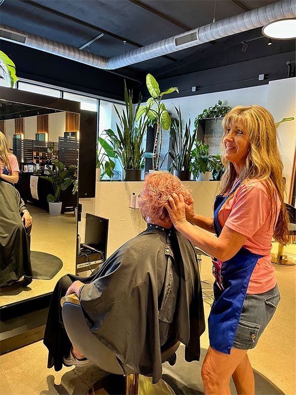 Woman styling client's hair in a salon. Both are smiling, plants and mirror visible.