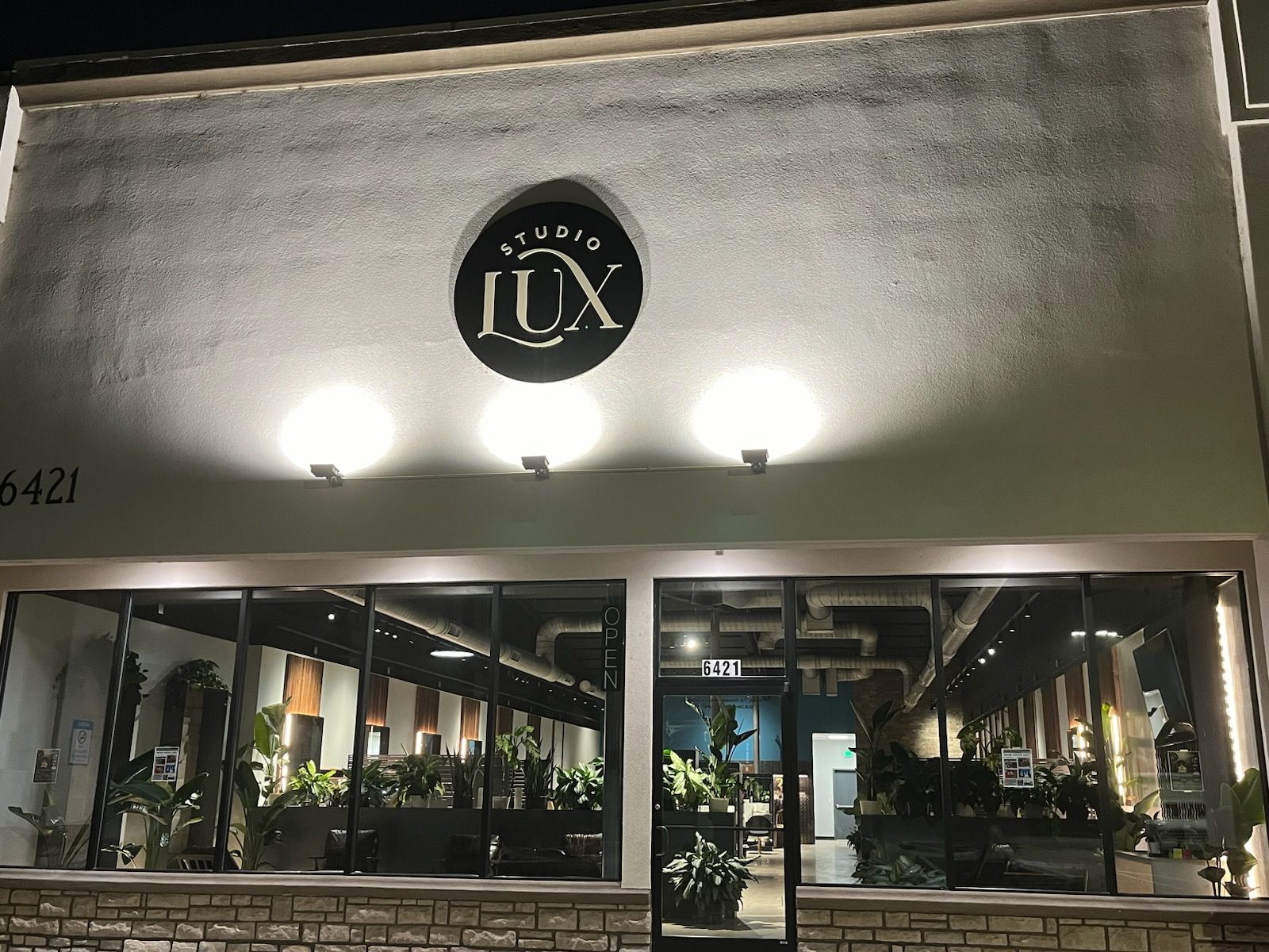 Studio LUX storefront, black sign, glass windows with plants, night.