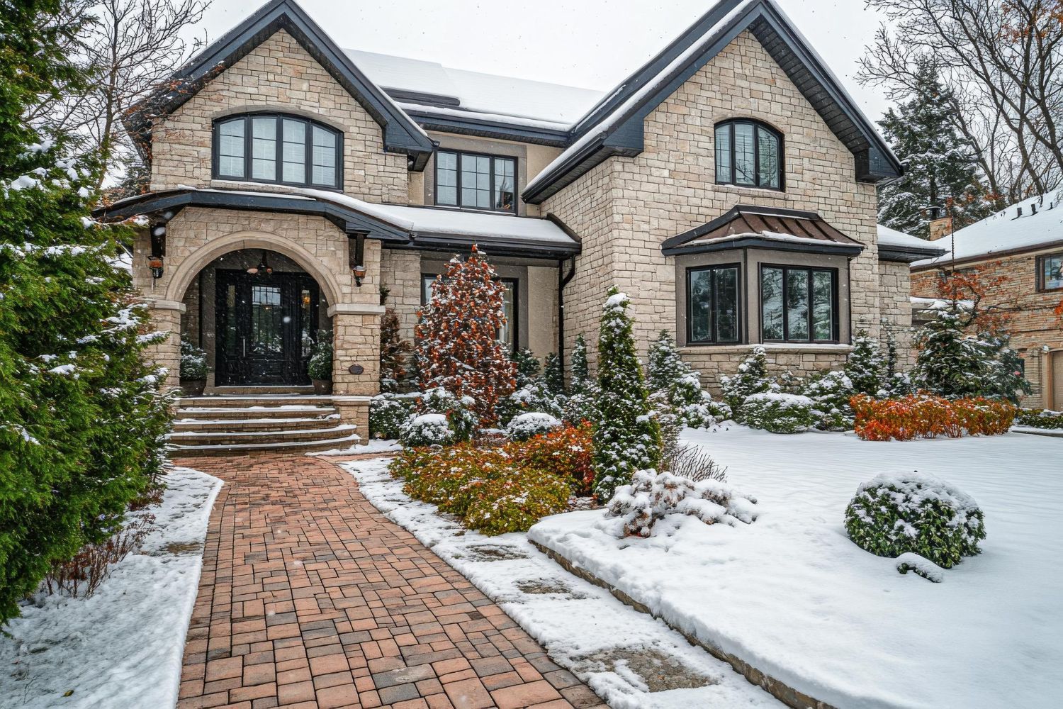 Snow-covered brick house with stone facade, arched entryway, and landscaping. A walkway leads to the front door.