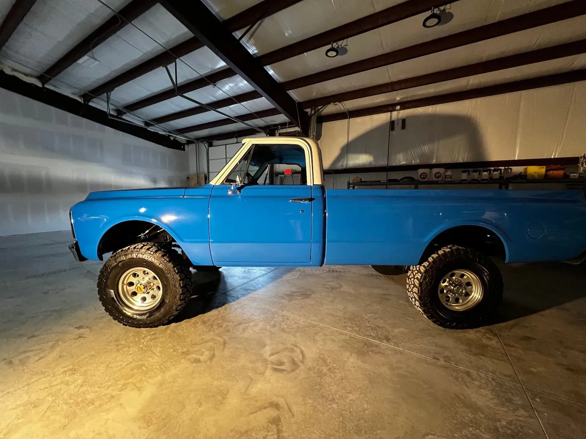 Blue lifted vintage pickup truck in a garage with a white cab, large tires, and exposed beams. | Gearhead Autoworks
