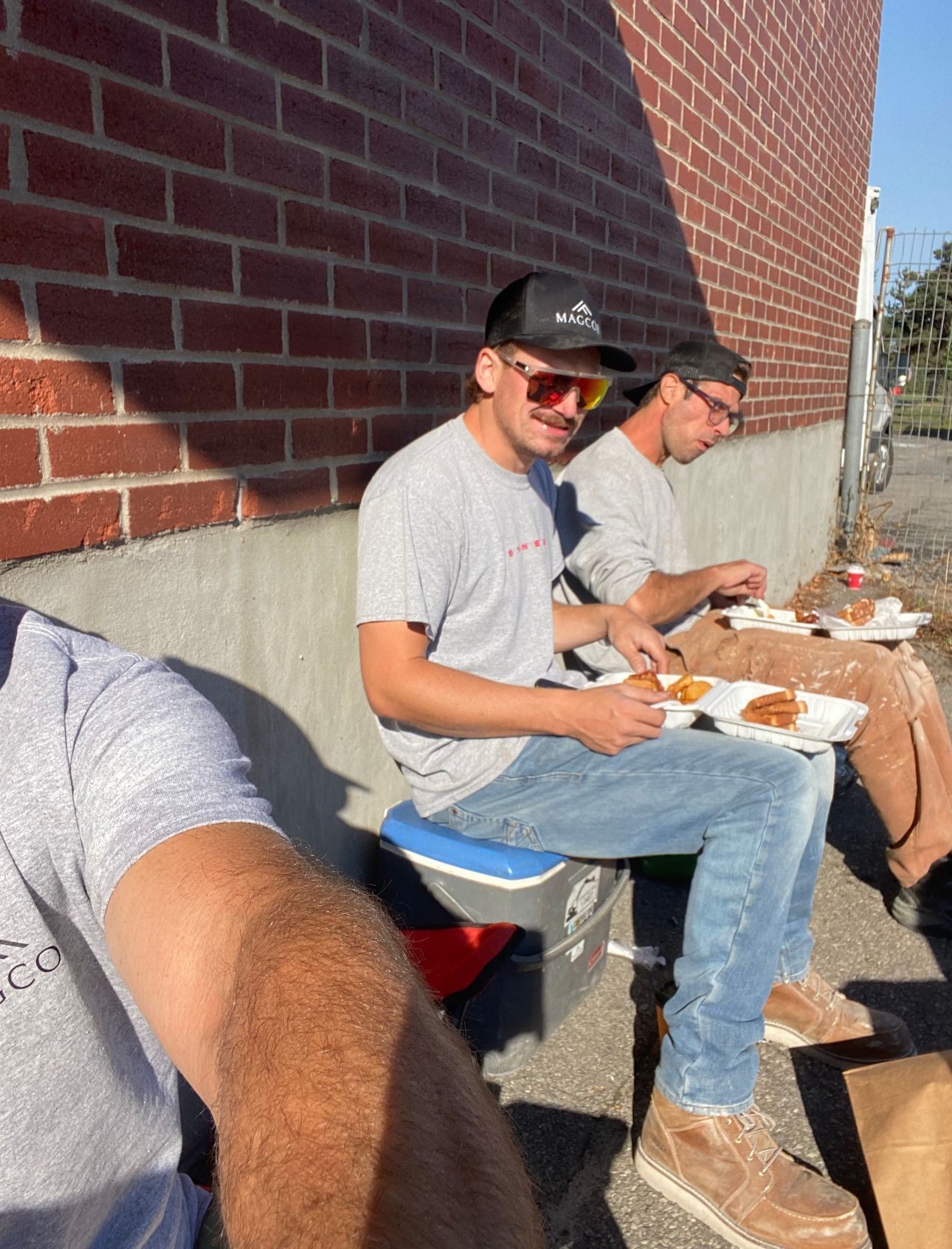 Two men are sitting on a cooler eating food in front of a brick wall.