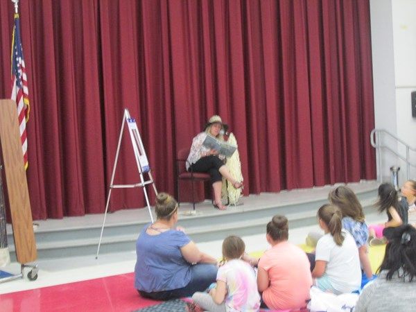 A woman is reading a book to a group of children in front of a red curtain.