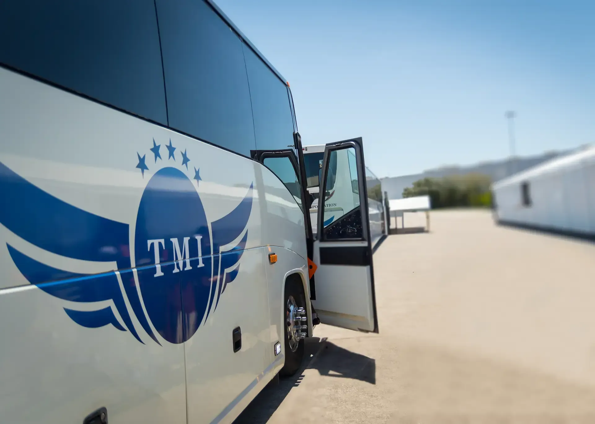 White tour bus with blue TMI logo parked in a lot, front door open under a clear sky.
