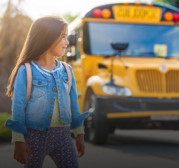 Girl with a backpack looking toward a yellow school bus on a sunny day.