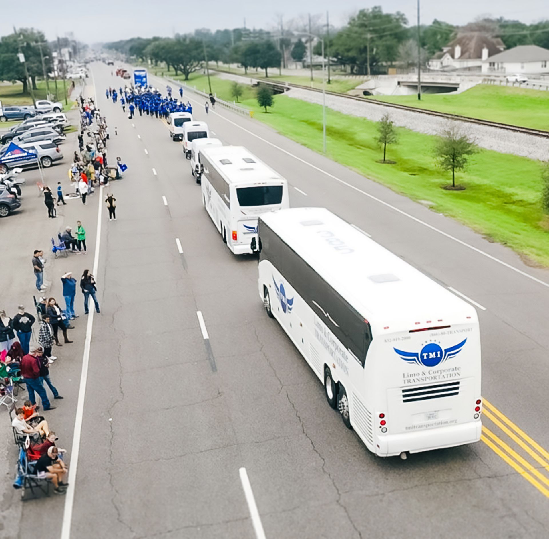 Buses and parade attendees lined up on a street with parked cars and a railroad in the background.