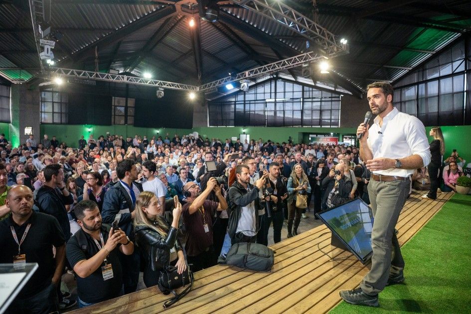 Man speaking at event, crowd watching; green accents, stage.
