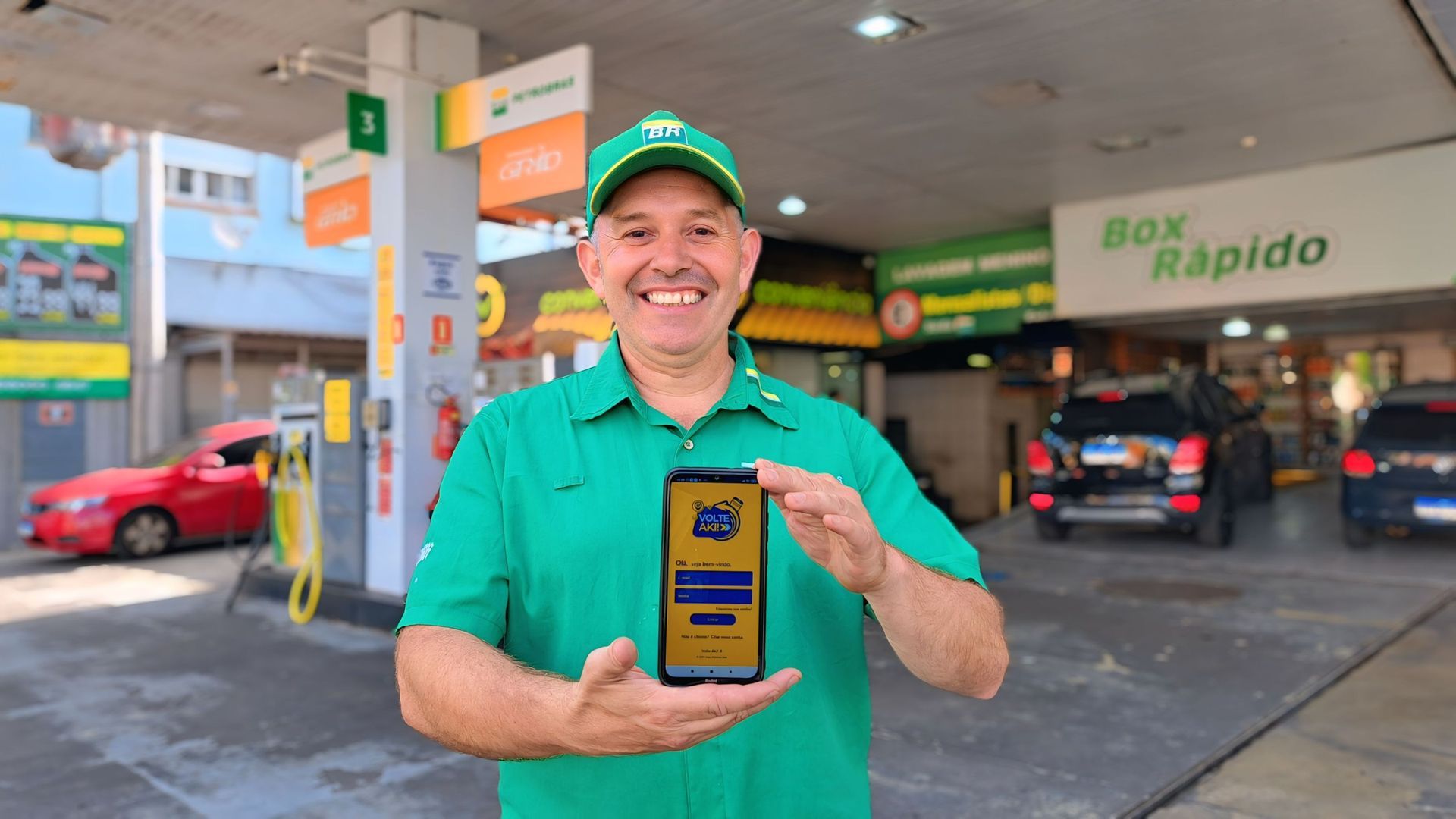 Homem de uniforme verde em um posto de gasolina sorri enquanto segura um celular com uma tela de aplicativo.