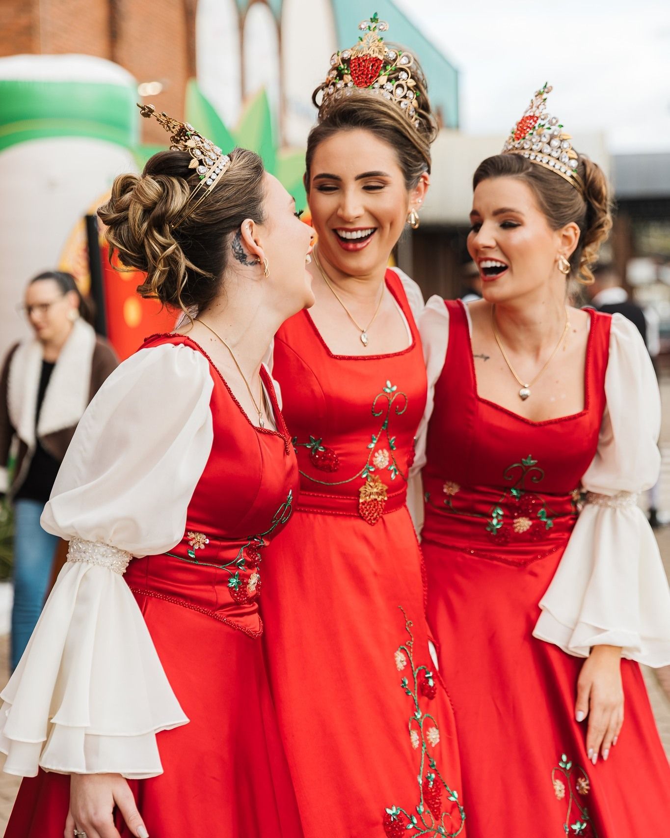 Três mulheres com vestidos vermelhos e coroas, rindo ao ar livre.