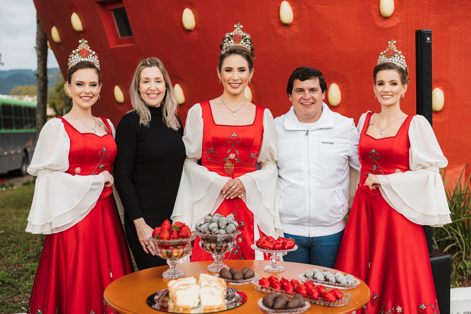 Pessoas posando em frente a um prédio em formato de morango. Mulheres em vestidos vermelhos com coroas e um homem.