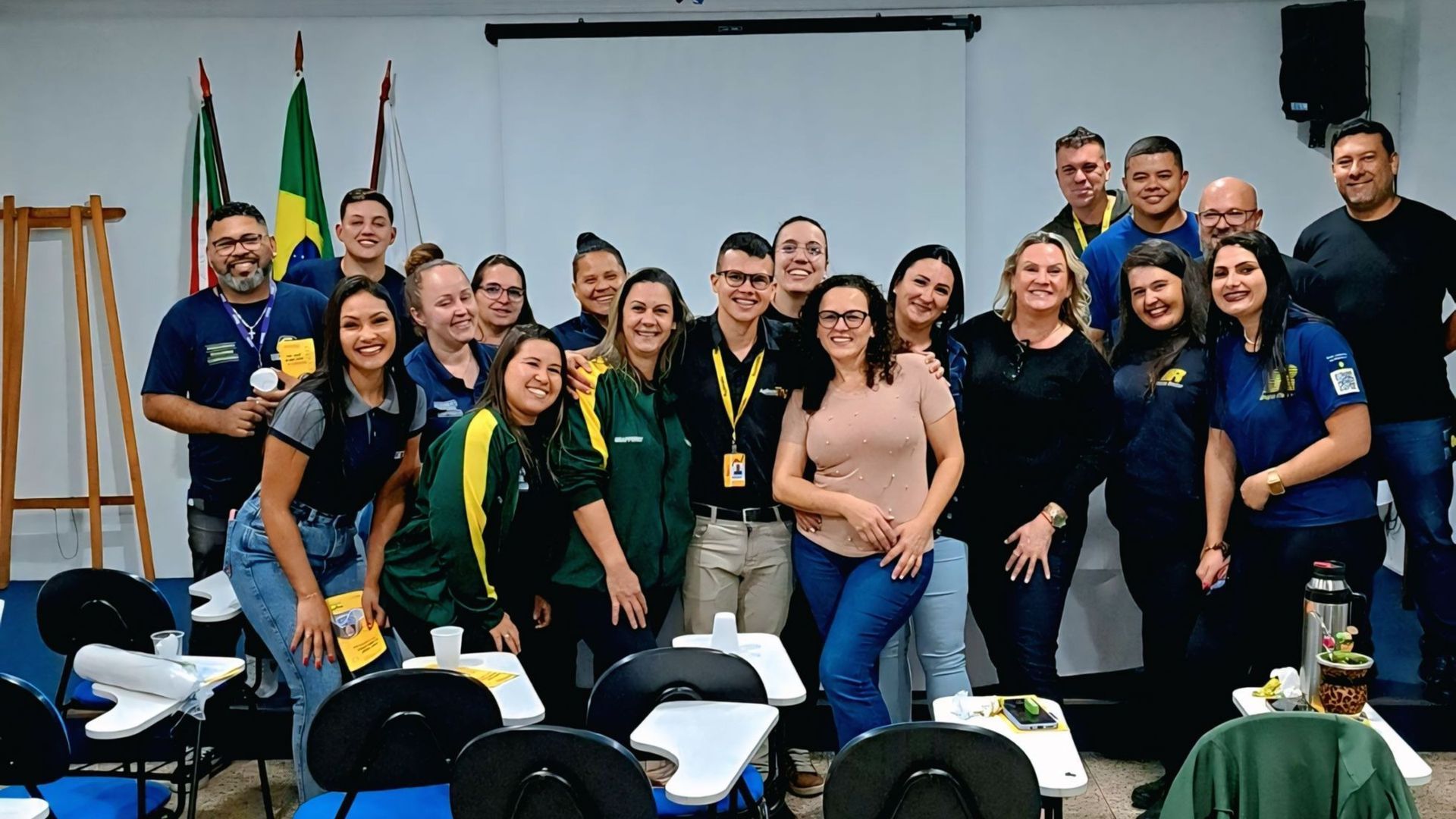 Foto de grupo: pessoas diversas sorrindo em uma sala com bandeiras do Brasil. Algumas seguram papéis, em um ambiente profissional.