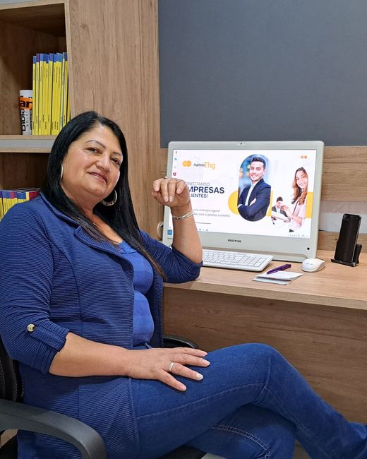 Mulher sentada à mesa com um computador exibindo um gráfico promocional. Ela está vestida de azul e sorrindo.