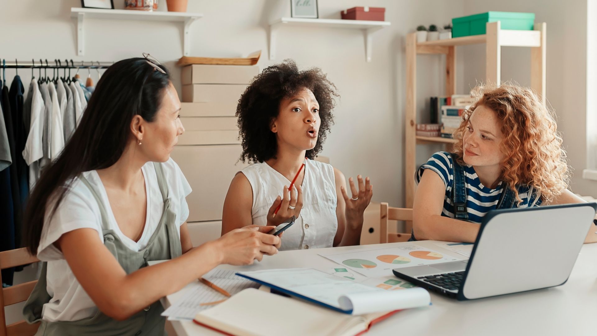 Three women in discussion around a table with laptop and papers, possibly planning in a light-filled room.