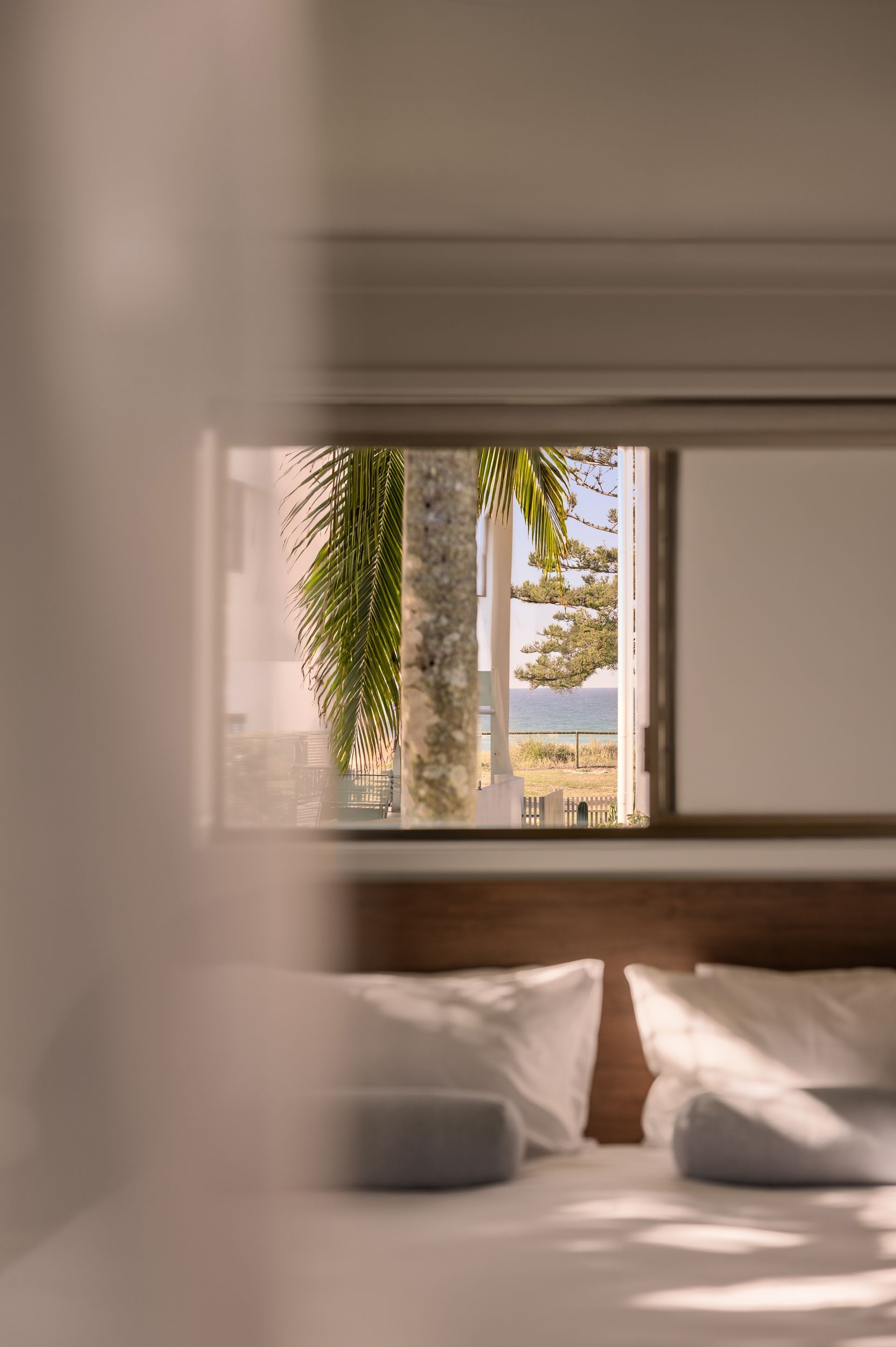 Sunlit bedroom with white bedding, viewed through a doorway toward trees and a distant ocean view