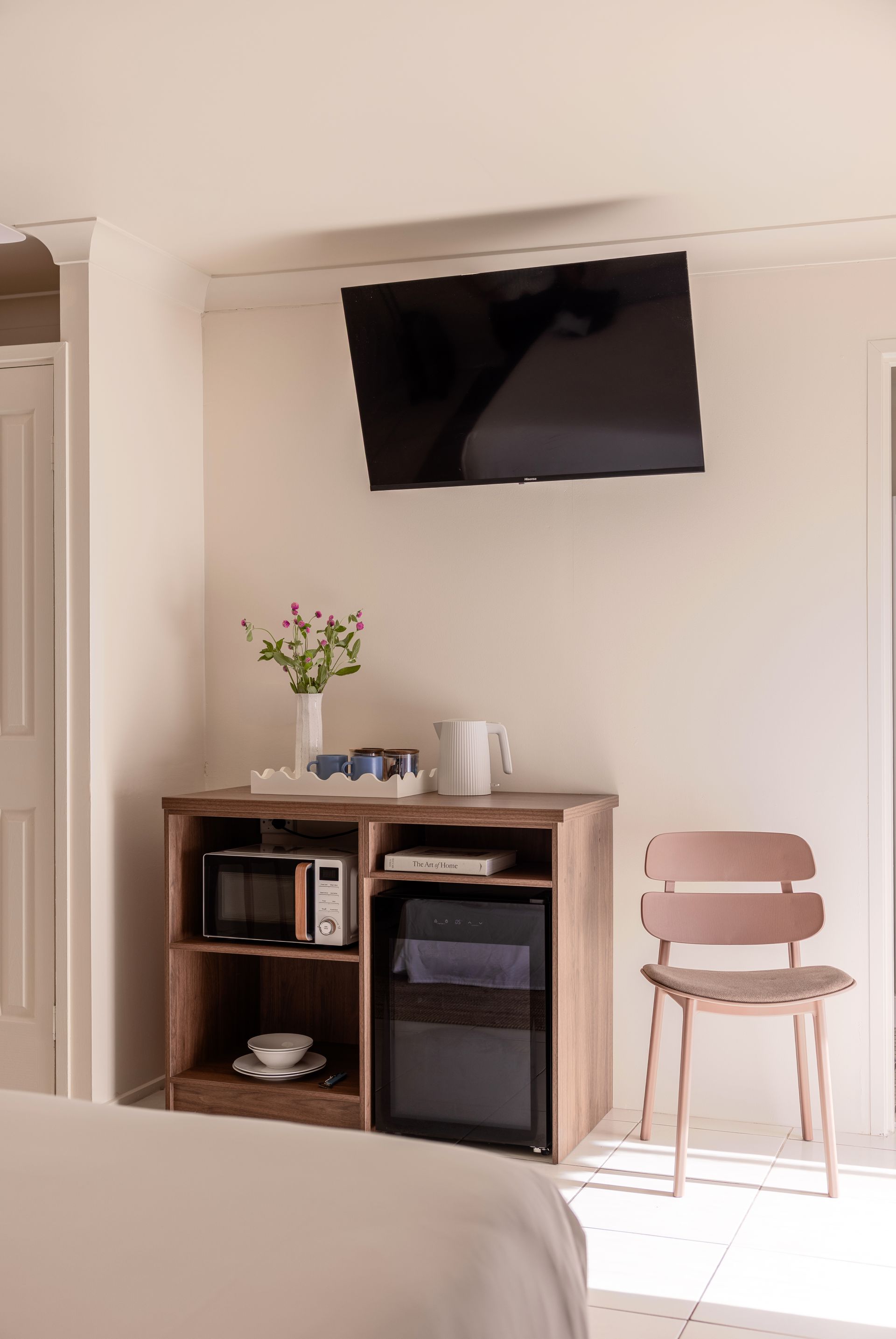 Small hotel room with wall-mounted TV, wooden minibar cabinet, and pink chair by a bright window.