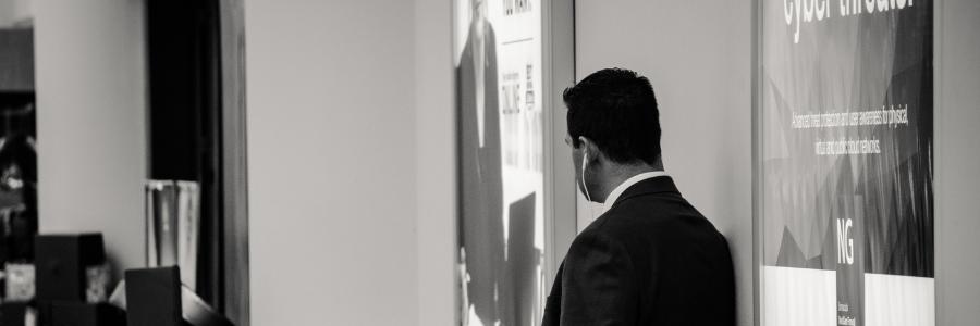 Man in suit standing in front of a poster, looking down, side profile. Black and white.