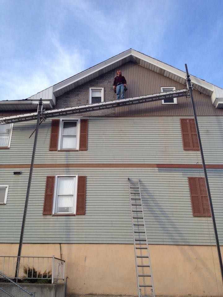 Person stands on scaffolding, working on a two-story house with green siding and brown shutters, cloudy sky.