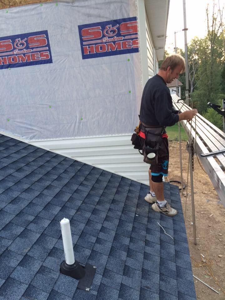 Roofer on a shingled roof, working near an unfinished wall. He's wearing a tool belt, knee brace. Sunlight is present.