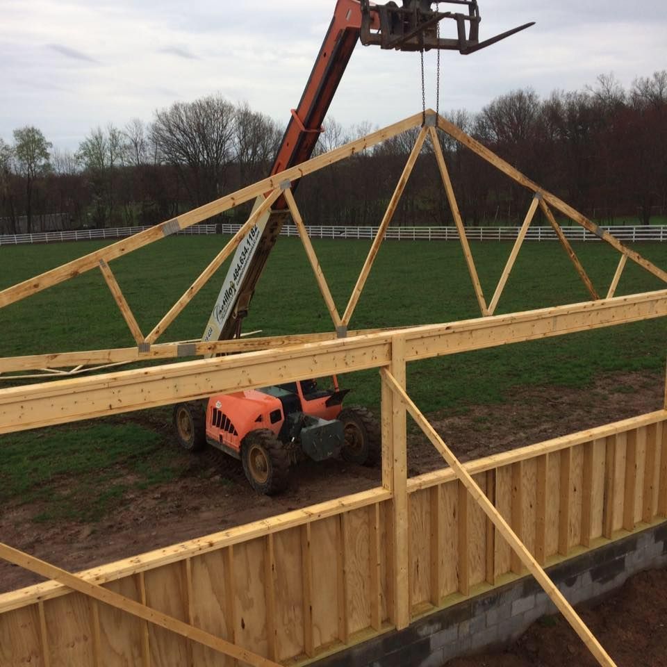 Orange forklift lifting a wooden roof truss onto a wood-framed building under construction, in a grassy field.