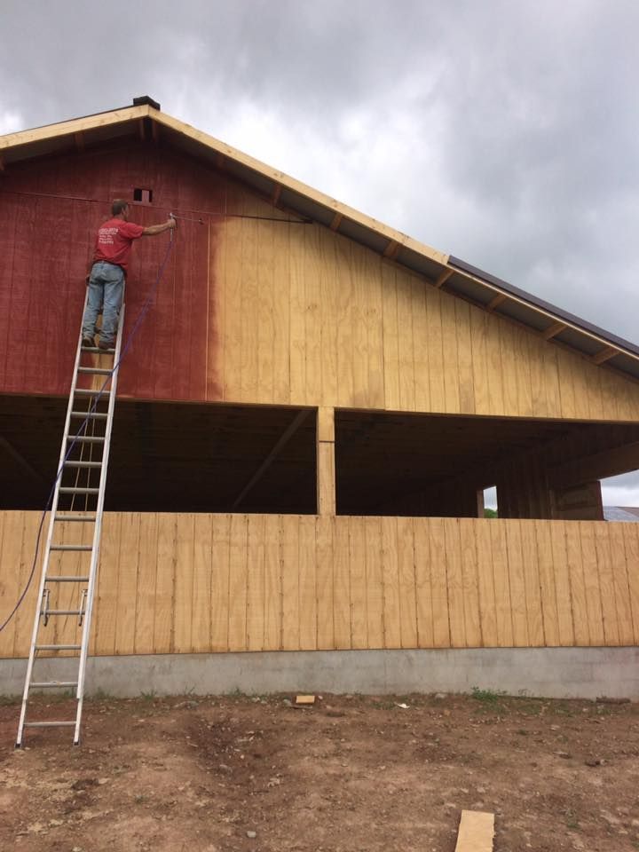 Person on a ladder painting a barn red. The unfinished barn is yellow with an open area. Cloudy sky.