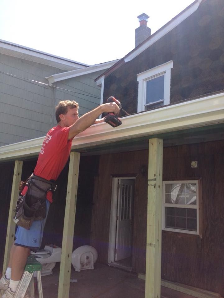 Man on a ladder installing a gutter with a power drill on a house.