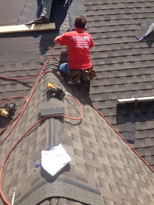 Roofer in red shirt kneels on a brown shingled roof, working with a hose and tools.