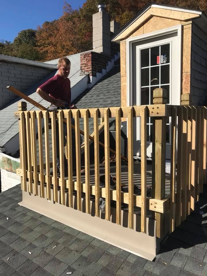 Man building wooden railing on rooftop, with dormer window in the background. Bright sunny day.