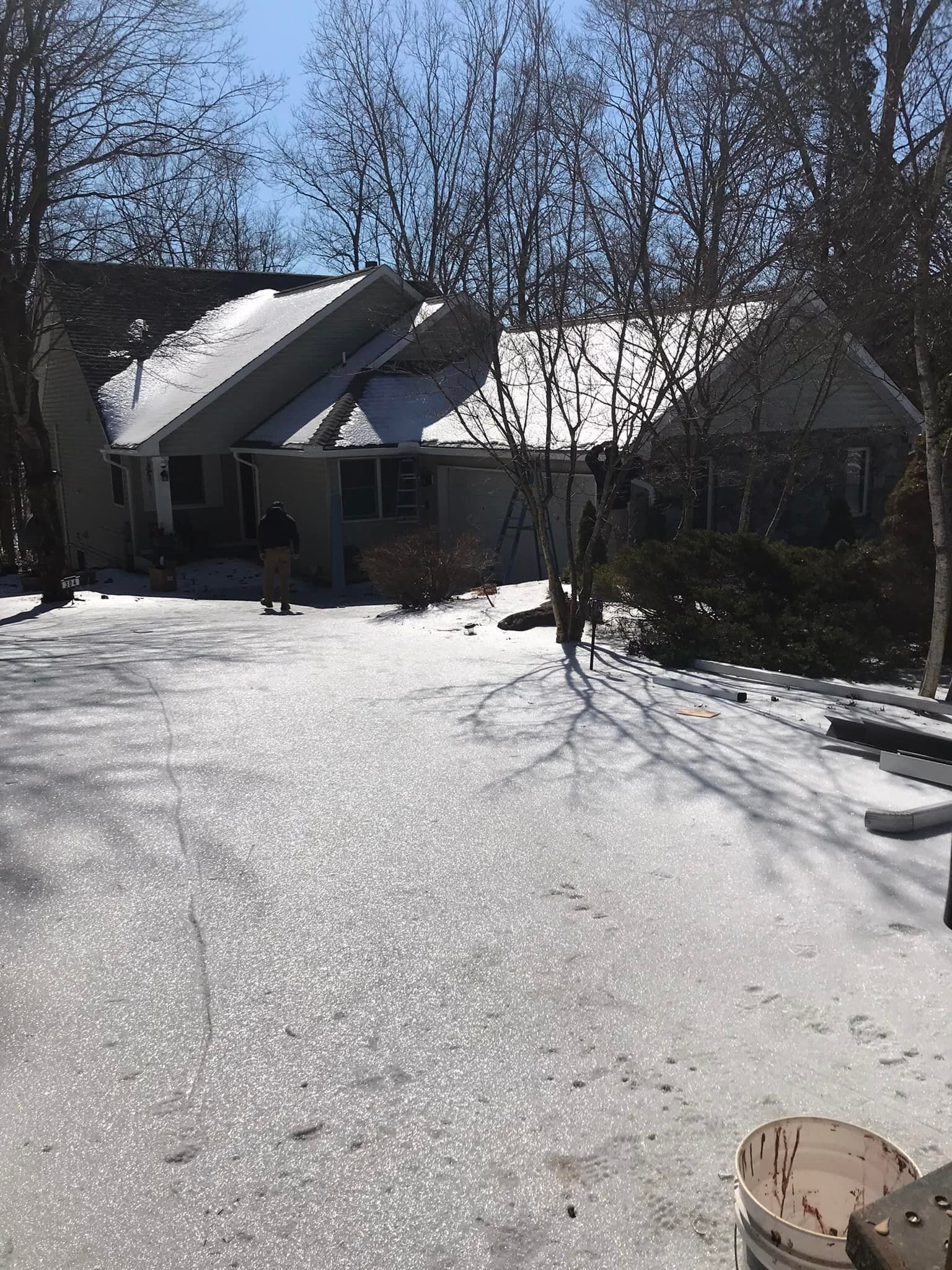 House with snowy roof and driveway; trees surround.