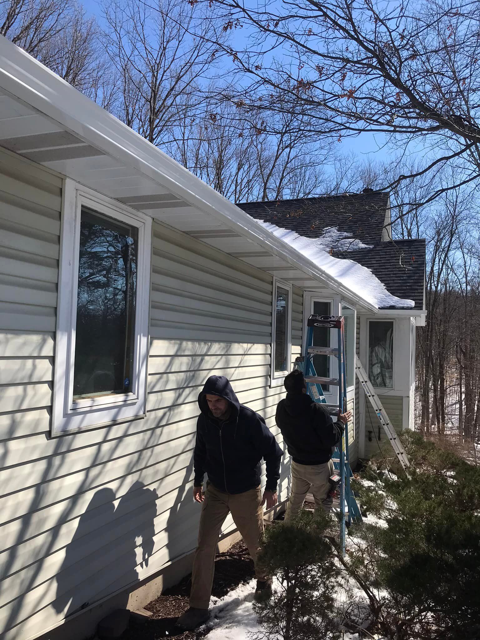 Two people working on a house with a ladder. Snow on roof. Sunny day.